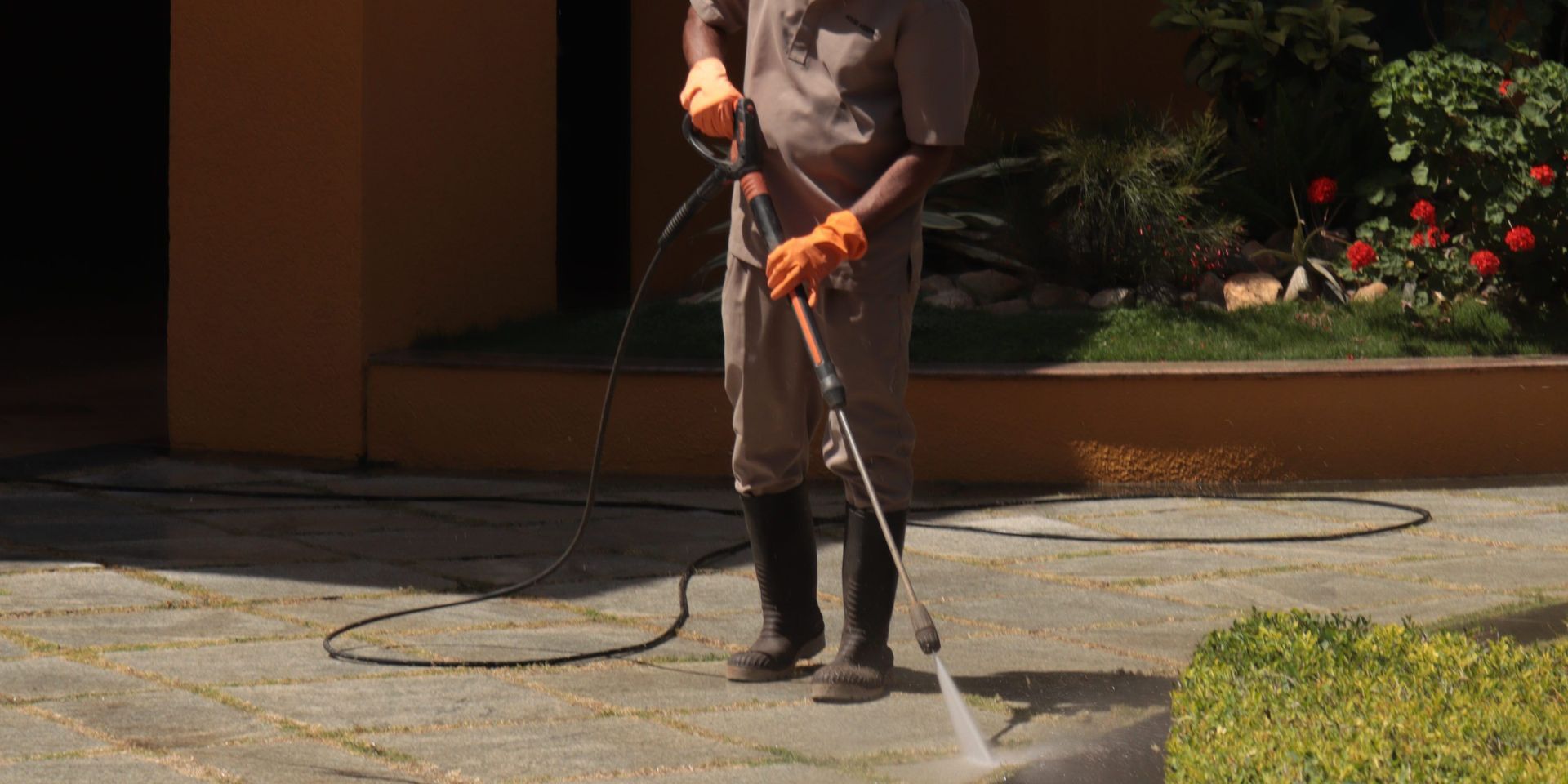 A gardener wearing a hat cleans an urban sidewalk with a pressure washer.